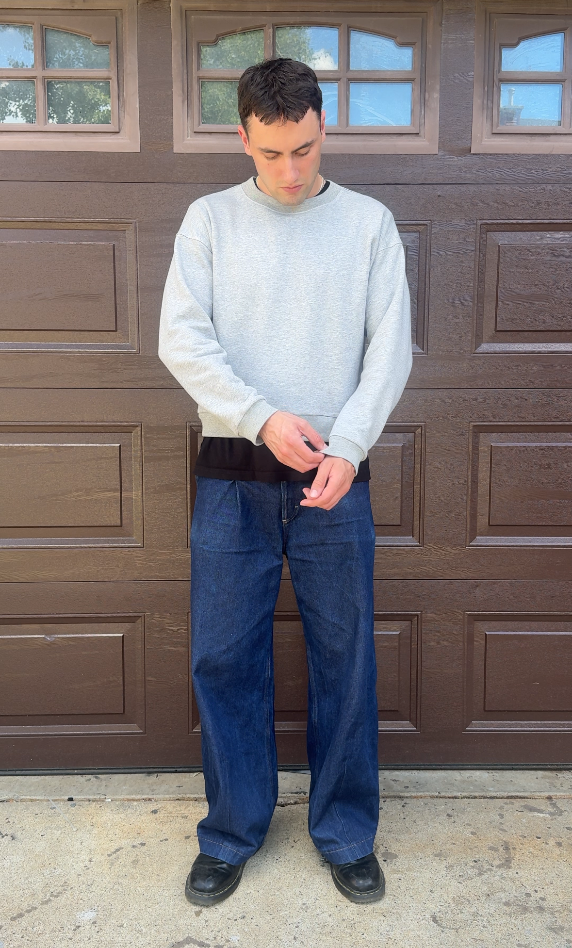 A man wearing a light gray long-sleeve mock neck sweater, loose dark blue jeans, and black shoes stands in front of a brown garage door.