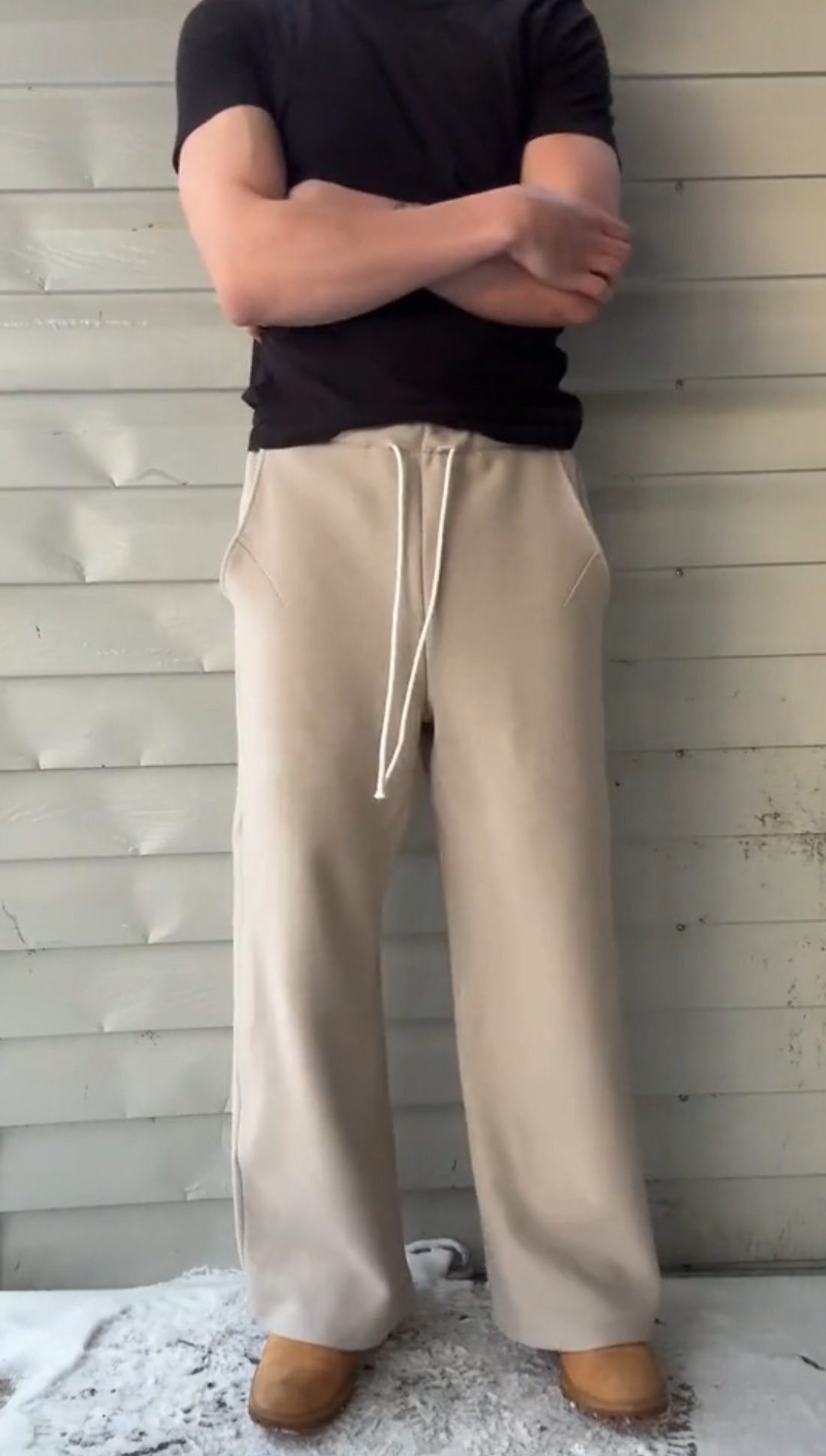 Person wearing a black shirt and beige pants standing against a light wooden panel wall.