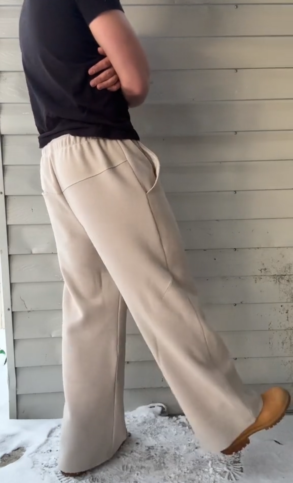 Person wearing beige wide-leg pants and a black top against a wooden panel background.